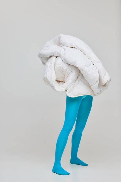 Portret van een jong meisje in een blauwe panty bedekt met een witte deken geïsoleerd over een grijze studio achtergrond. Ik voel me warm en veilig. Concept van retro mode, kunst fotografie, stijl, queer, schoonheid, advertentie - Foto, afbeelding