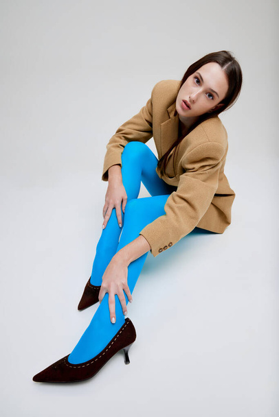 Portrait of young beautiful girl posing in blue tights, oversized jacket and heels isolated on grey studio background. Fashionable. Concept of modern fashion, art photography, style, queer, beauty, ad - Foto, imagen