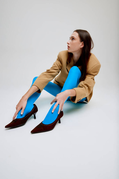 Young beautiful girl sitting on floor, posing in blue tights, oversized jacket and heels isolated on grey studio background. Youth. Concept of modern fashion, art photography, style, queer, beauty, ad - Фото, зображення
