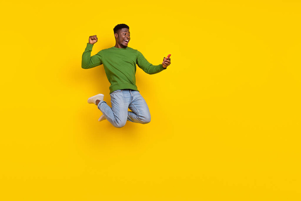 Full length body size view of handsome trendy guy using device jumping having fun isolated over vivid yellow color background. - Фото, изображение