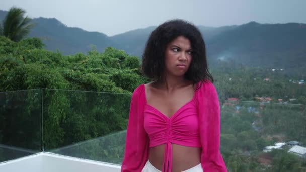Egy unatkozó fiatal nő portréja, aki reménytelenül és kétségbeesetten néz a kamerába, az erkélyen, kilátással egy trópusi falura. - Felvétel, videó