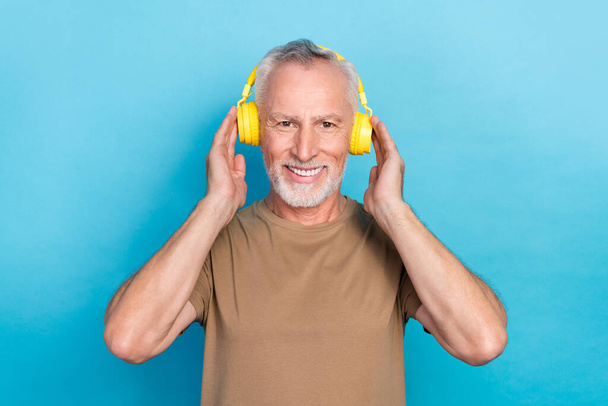 Photo of cheerful glad retired man gray hairstyle stubble wear beige t-shirt hands touch headphones isolated on blue color background. - Zdjęcie, obraz