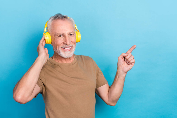 Portrait of positive senior gray hair beard dressed beige t-shirt headphones indicating empty space sale isolated on blue color background. - Фото, изображение