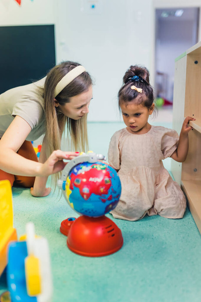 Die blonde Lehrerin berührt die kleine Plastikkugel und spricht mit einem kleinen Mädchen im Kinderzimmer, vertikale Aufnahme. Hochwertiges Foto - Foto, Bild