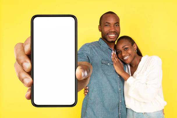 Boldog fekete házastársak mutatja Big Phone üres képernyős mosolygós kamera ajánló új App pózol ölelés sárga háttér stúdióban. Mobil alkalmazás koncepció. Mockup - Fotó, kép