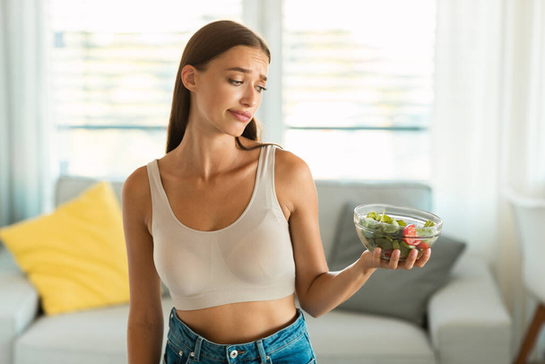 Gewichtsverlust. Unzufriedene junge Frau mit Blick auf Gemüsesalat Schlankheitswahn posiert zu Hause. Konzept für gesunde Ernährung und Ernährungsprobleme - Foto, Bild