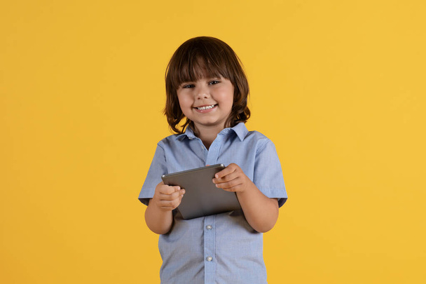 Modern çocuk eğlencesi. Dijital tablet tutan mutlu küçük çocuğun stüdyo portresi ve kameraya gülümsemesi, turuncu arka plan, serbest alan - Fotoğraf, Görsel