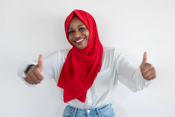 Líbí se mi to a doporučuji to. Šťastná černá muslimka v hidžábu gestikuluje palci nahoru a usmívá se na kameru, pózuje na pozadí bílé studio zdi. Koncept gesta palce nahoru - Fotografie, Obrázek
