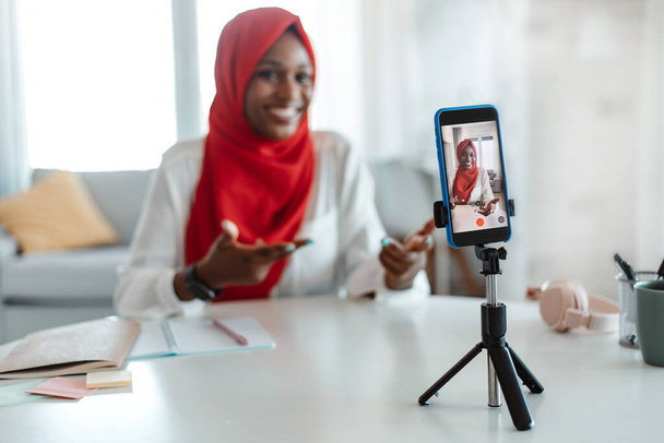 Θετική μαύρη ισλαμική κυρία σύλληψη περιεχομένου για vlog στο σπίτι, εγγραφή βίντεο σε smartphone σε τρίποδο, ομιλία και χειρονομίες στην κάμερα της συσκευής, επιλεκτική εστίαση - Φωτογραφία, εικόνα