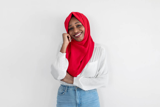 Улыбающаяся чёрная мусульманка в хиджабе позирует над белой стеной студии, смотрит в камеру, свободное место. Веселая африканская американка в красном платке. Концепция мусульманской красоты - Фото, изображение
