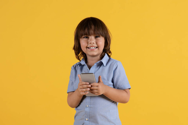Kids and gadgets. Adorable little boy holding smartphone and smiling to camera, watching videos online, orange studio background, free space - Photo, Image