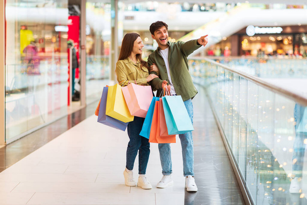 Cheerful Couple Shopping Pointing Finger Aside Advertising Great Black Friday Sale Offer Posing Holding Shopper Bags Standing In Mall Indoors. Sales Season And Commerce Concept - Photo, Image