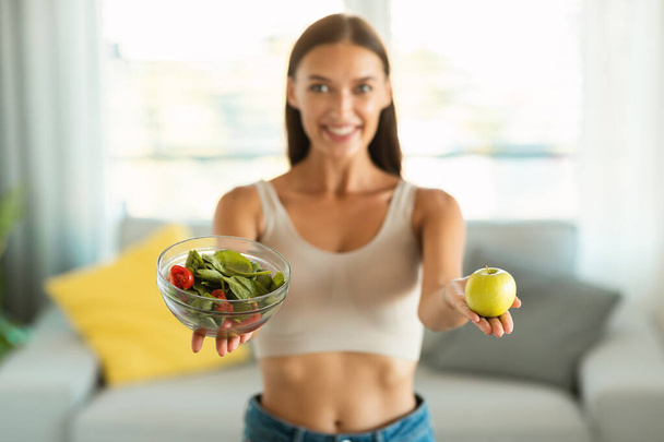 Gesunde Ernährung. Glückliche Frau zeigt Salat in der Schüssel und Apfelfrüchte, die eine Gewichtsabnahme empfehlen und lächelt lächelnd in die Kamera. Schlankheitskonzept. Selektiver Fokus auf Lebensmittel - Foto, Bild