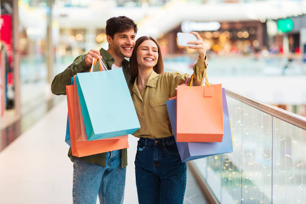 Προσφορά πωλήσεων. Ευτυχισμένοι πελάτες ζευγάρι ψώνια και κάνοντας Selfie στο κινητό τηλέφωνο διασκεδάζοντας στο σύγχρονο εμπορικό κέντρο εσωτερική. Αγοραστές λήψη φωτογραφιών στο τηλέφωνο απόσπαση με τσάντες Shopper. Έννοια Shopaholism - Φωτογραφία, εικόνα