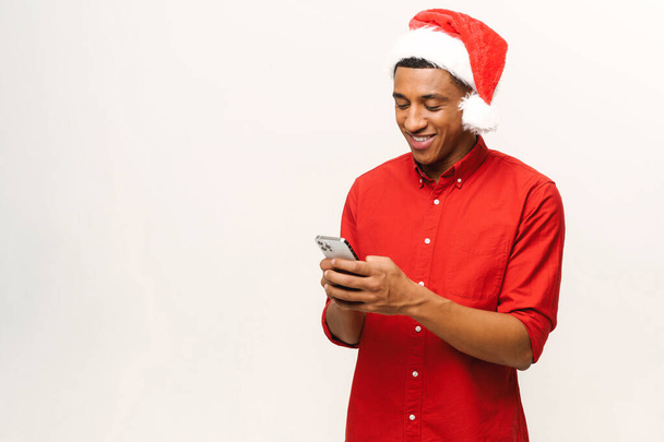 Cheerful attractive young man in santa hat holding smartphone, enjoying chatting in social networks, texting messages with congratulations, smiling, using mobile app, isolated on white, copy space - Zdjęcie, obraz