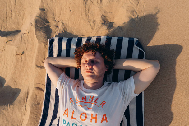 Молодой кавказский мужчина с вьющимися волосами, закрытыми глазами, отдыхающий, расслабляющий или спящий в белой футболке со словами Саммер Алоха лежит на полосатом полотенце на песчаном пляже бархан или песчаная дюна на закате. Портрет - Фото, изображение