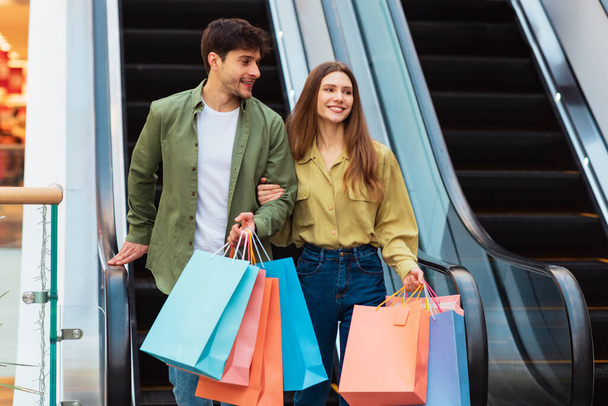 Fekete Péntek Ajánlat. Happy Customers Couple Shopping Hordozó Shopper Bags Walking Near Moving Stairs In Modern Mall. Weekend Sales Advertisement, Fogyasztói koncepció - Fotó, kép