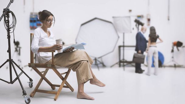 Professional actress sitting on the director's chair and reading the script, film studio interior in the background: film industry and video production concept - Foto, Imagem