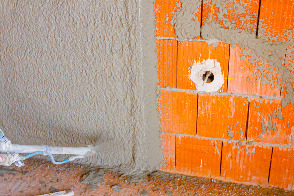 Worker man is holding mortar hose in his hand and plastering under pressure interior brick walls with one exposed electrical socket with cable, building under construction. - Foto, Imagen