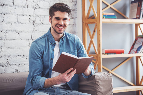 attraktiver junger Mann liest ein Buch, blickt in die Kamera und lächelt, während er zu Hause auf dem Sofa sitzt - Foto, Bild