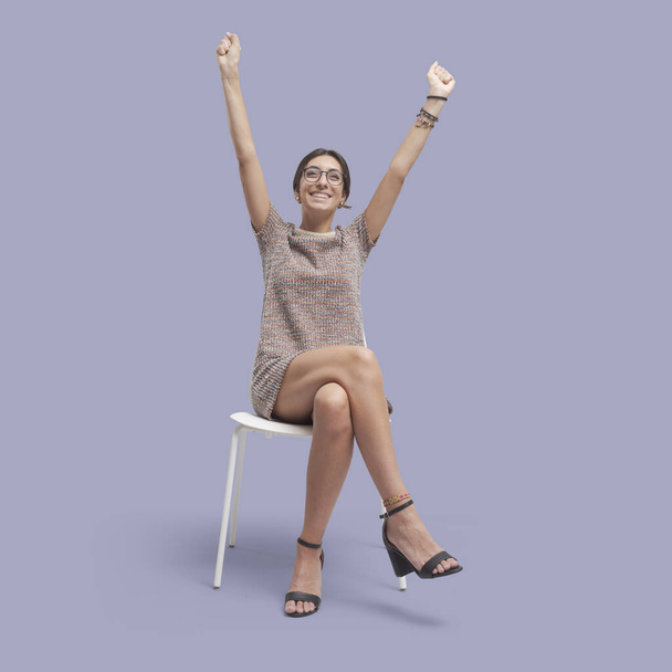 Cheerful woman sitting on a chair and celebrating with raised arms - Fotoğraf, Görsel