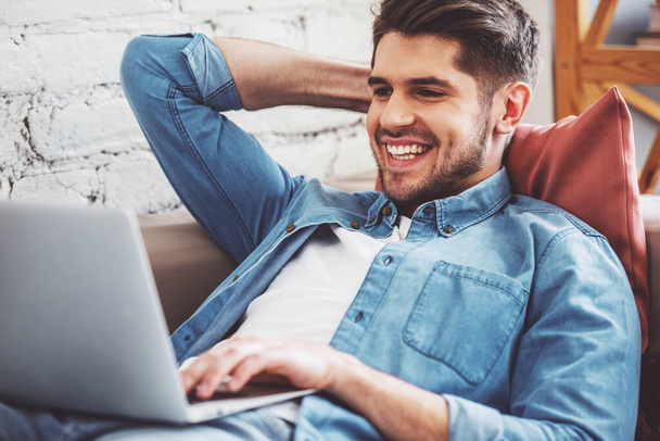 Joven atractivo está utilizando un ordenador portátil y sonriendo mientras está acostado en el sofá en casa
 - Foto, imagen