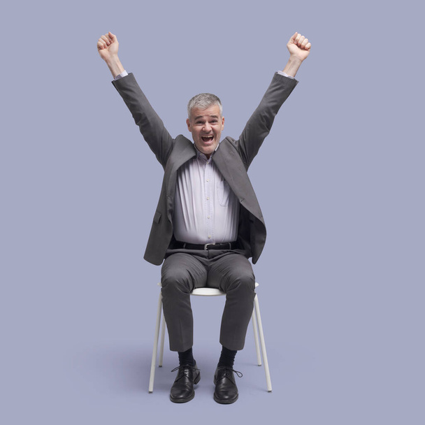 Onnellinen menestynyt mies istuu tuolilla ja juhlii kädet ylhäällä - Valokuva, kuva