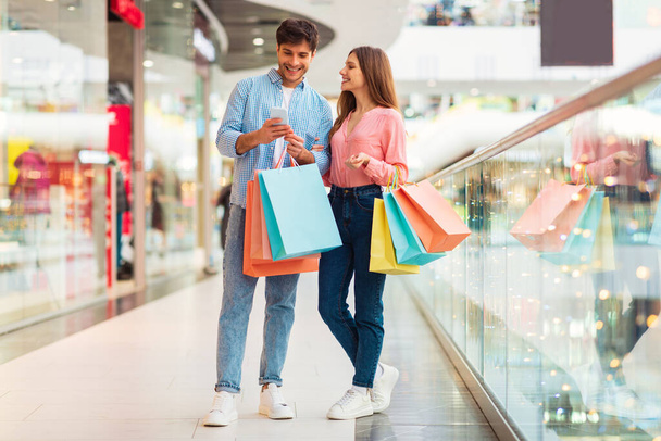 Cheerful Spouses Shopping Using Mobile Phone Posing Holding Colorful Shopper Bags In Modern Mall Indoors. Buyers Couple Purchasing Clothes Online Via Application. Sales Concept. Full Length - Photo, Image