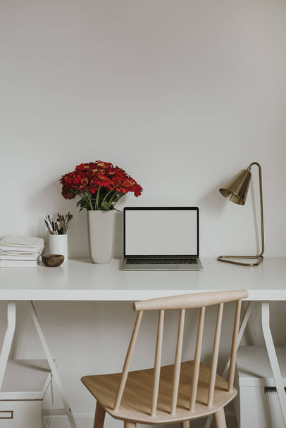 Computer portatile con schermo bianco sul tavolo con bouquet di fiori di gerbero rosso. Modello di interior design per spazi di lavoro in stile influencer estetico con spazio copia mockup - Foto, immagini