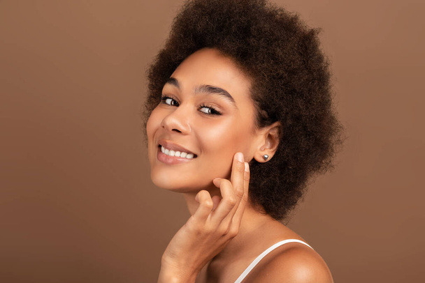 Smiling young mixed race female with curly hair applies cream on face, touches perfect skin with fingers isolated on brown background, close up. Natural beauty care, spa treatment and lifestyle - Foto, afbeelding