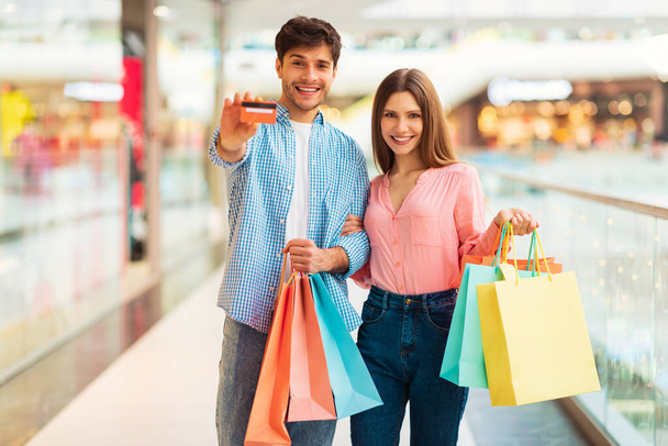 Tolle Verkäufe. Fröhliche Ehepartner zeigen Kreditkartenbesitzern Papiertaschen, die lächelnd vor der Kamera in einem modernen Einkaufszentrum posieren. Black Friday Angebotskonzept. Selektiver Fokus - Foto, Bild