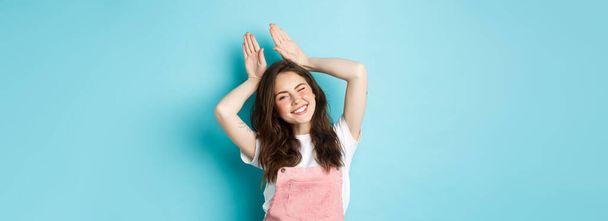 Cheerful girl celebrating Easter, showing easter bunny gesture and smiling cute at camera, standing over blue background in spring outfit. - Photo, Image