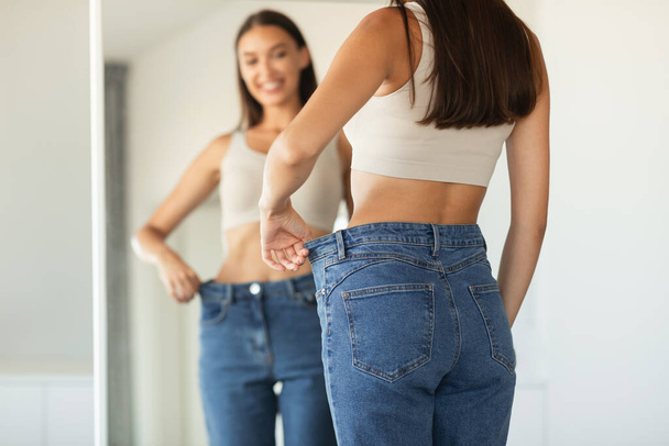 Gewichtsverlust. Lady trägt übergroße Jeans im Größenvergleich nach dem Abnehmen posiert in der Nähe von Spiegel zu Hause. Selektiver Fokus auf den weiblichen Rücken. Abgeschnittener Schuss, flache Tiefe - Foto, Bild