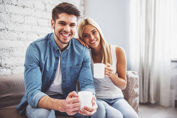Beautiful young couple is holding cups of drink, looking at camera and smiling while sitting on sofa at home - Photo, Image