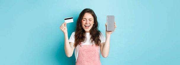 Excited attractive woman scream of joy, showing empty smartphone screen and plastic credit card, raising hands up and smiling happy, standing over blue background. - Photo, Image