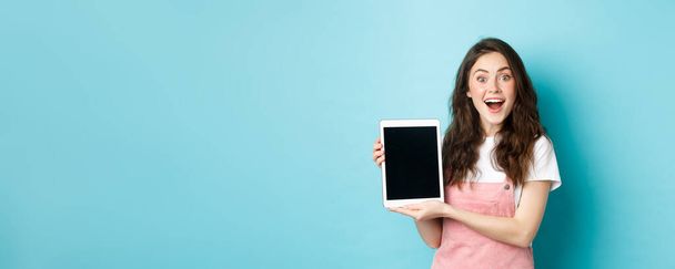 Amüsiert hübsche Mädchen zeigen leeren digitalen Tablet-Bildschirm und lächelt aufgeregt in die Kamera, zeigt ehrfürchtige Promo-Angebot, vor blauem Hintergrund stehen. - Foto, Bild