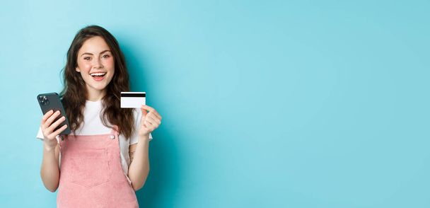 Tyylikäs nuori nainen verkkokaupoissa, näyttää muovinen luottokortti ja tilalla älypuhelin, seisoo sinistä taustaa vasten. - Valokuva, kuva