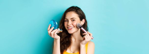 Beauty. Close up of attractive woman looking in mirror and using brush to apply make up, smiling pleased at camera, standing against blue background. - Fotoğraf, Görsel