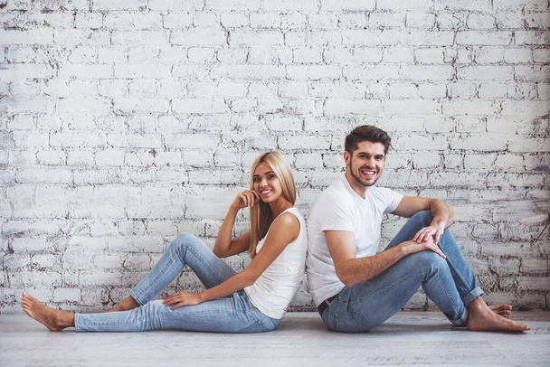 Beautiful young couple is looking at camera and smiling, sitting back to back on floor against white brick wall - Фото, изображение