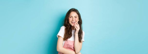 Portret szczęśliwej atrakcyjnej kobiety śmiejącej się i bawiącej, uśmiechającej białymi zębami, cieszącej się zabawnym filmem, stojącej na niebieskim tle. - Zdjęcie, obraz