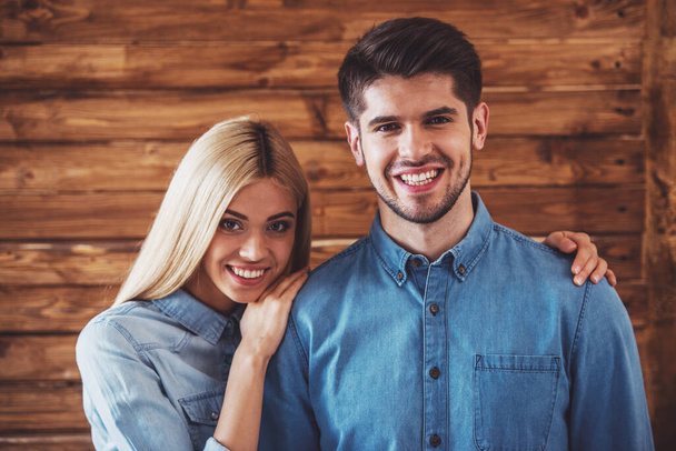 Portret van mooie jonge paar in jeans kleding knuffelen, camera kijken en glimlachend, permanent tegen houten muur - Foto, afbeelding