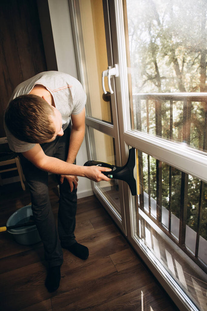 Ένας άντρας πλένει παράθυρα στο σπίτι χρησιμοποιώντας μια ασύρματη ηλεκτρική σκούπα. Εμπρόσθια όψη, επιλεκτική εστίαση - Φωτογραφία, εικόνα