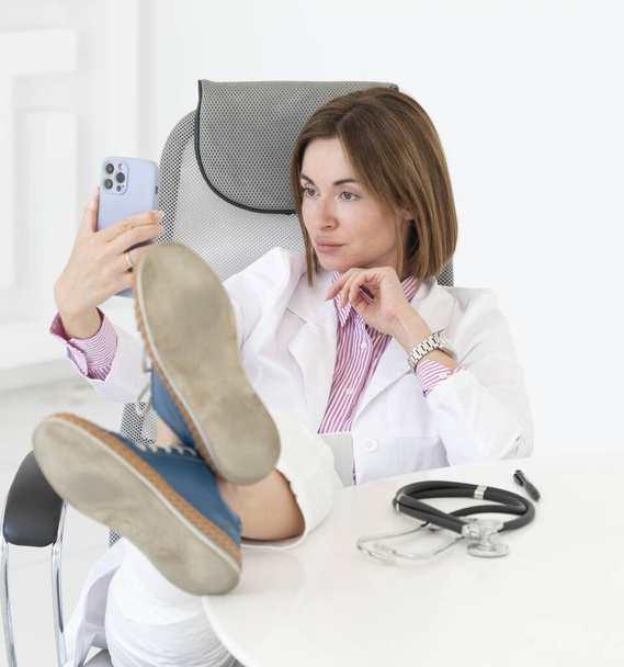 Молодая женщина-врач с ногами на рабочем месте расслабляет и фотографируется со смартфоном вместо работы - Фото, изображение