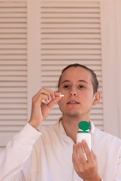 Vertical view of young man about to take a pill from a white and green container for medical purpose in white background - Foto, Bild