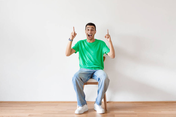 Overjoyed arab man sitting on chair and pointing two fingers up, offering space for your advertisement on light studio wall, full length. Best promotion, cool discount, huge sale concept - Photo, Image