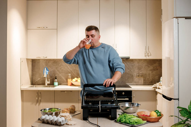 Ο άνθρωπος ετοιμάζει νόστιμο ζουμερό κρέας μπριζόλα σε μια ηλεκτρική ψησταριά σε ξύλινο τραπέζι. Καπνός στην κουζίνα του σπιτιού - Φωτογραφία, εικόνα