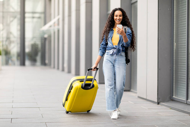 Cheerful pretty curly middle eastern young woman in comfy casual outfit going vacation alone, walking by airport with yellow luggage, using cell phone, online check-in, copy space, full length - Φωτογραφία, εικόνα