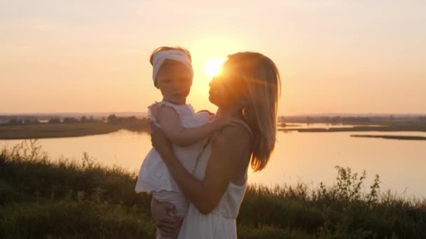 A woman holds her little daughter in her hands at sunset and kisses her on the cheek. Mid shot - Footage, Video