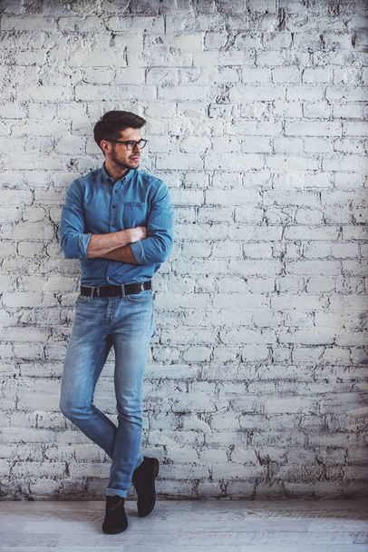 Retrato completo de un joven guapo vestido con jeans y anteojos mirando hacia otro lado y sonriendo, de pie contra la pared de ladrillo blanco
 - Foto, imagen