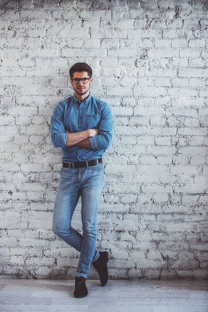 Full lenght portrait of handsome young man in jeans clothes and eyeglasses looking at camera and smiling, standing against white brick wall - Photo, Image
